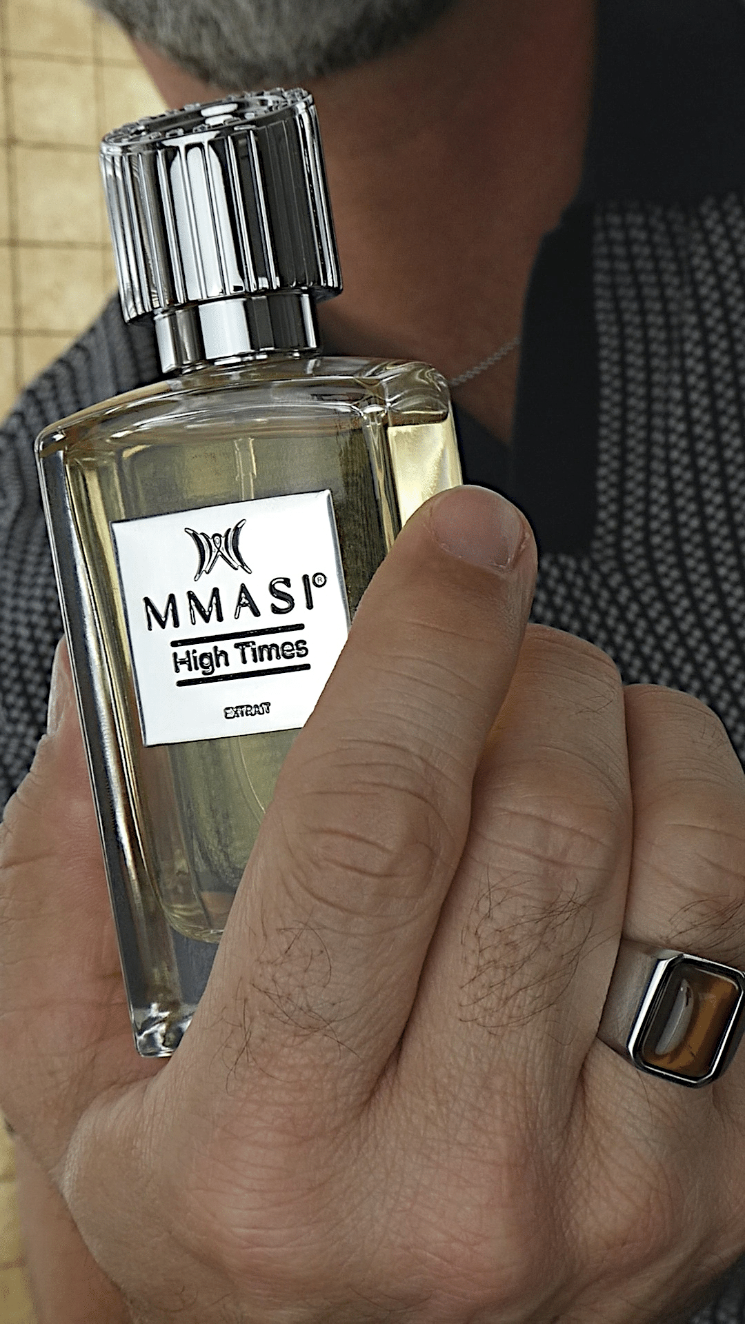 Close-up of a man's hand holding a rectangular bottle of MMASI High Times perfume with a silver cap and a silver and amber ring on his finger.