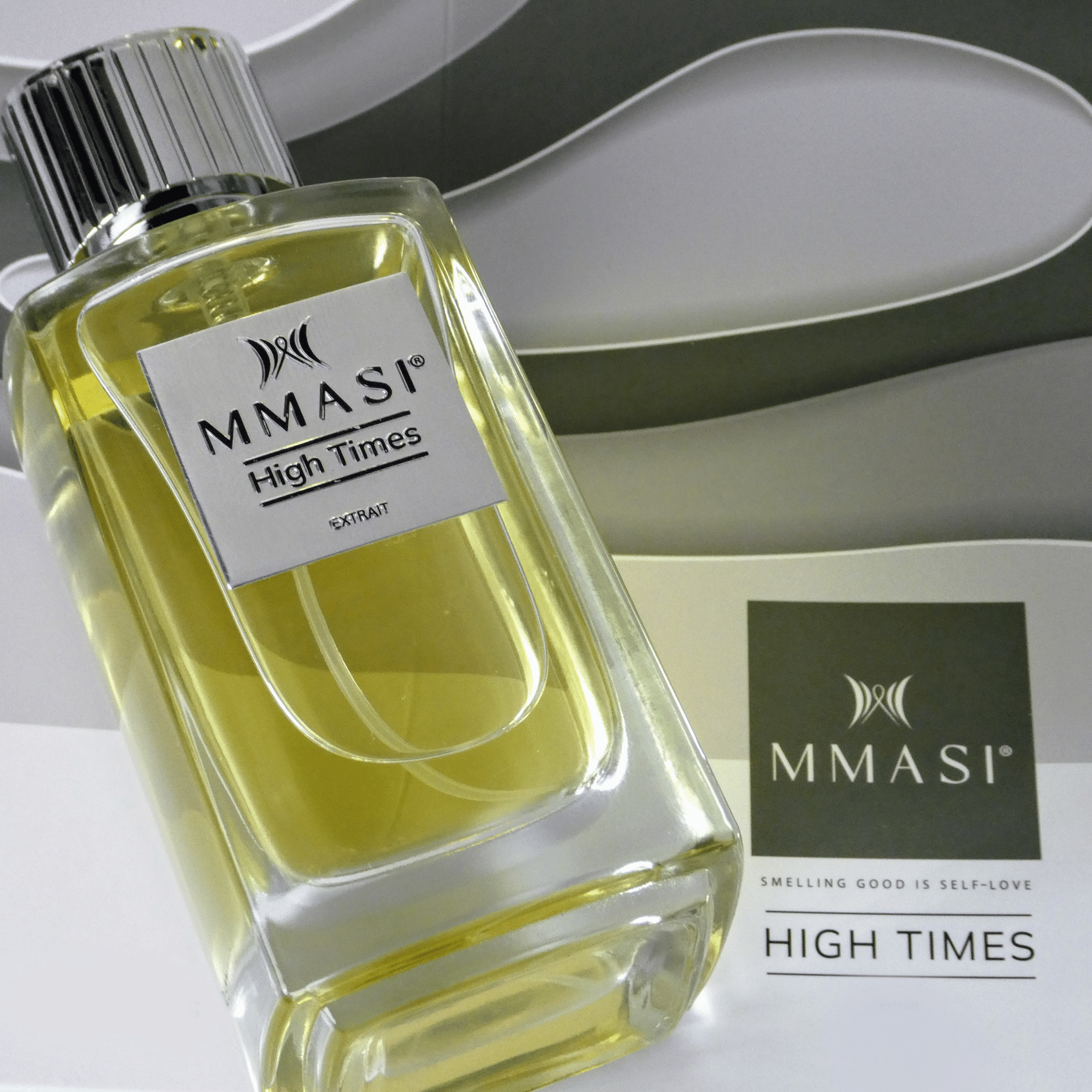 Close-up of a clear glass perfume bottle with yellow liquid, labeled MMASI High Times Extrait. Behind it is a gray and white curved pattern and a square with the MMASI logo and the tagline "Smelling good is self-love."