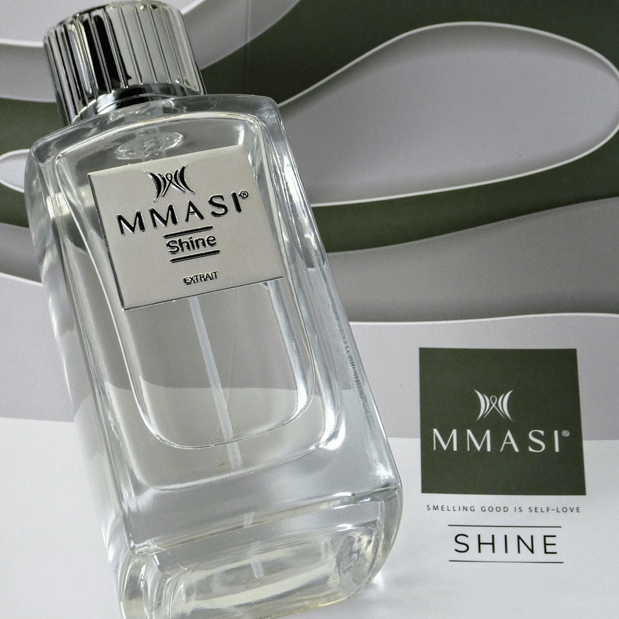 A clear bottle of MMASI Shine perfume with silver cap, placed diagonally in front of a gray and white background with a stylized M logo, accompanied by the tagline "SMELLING GOOD IS SELF-LOVE."