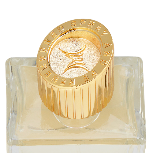 A gold circular perfume cap with the words “Spray and be remembered” embossed around the edge and an M stylized logo in the center, sitting on a clear rectangular base bottle