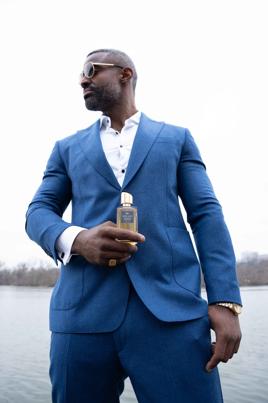 A stylish individual in a sharp blue suit holding a bottle of MMASI perfume, with a gold watch on the left wrist, against a natural water body backdrop.