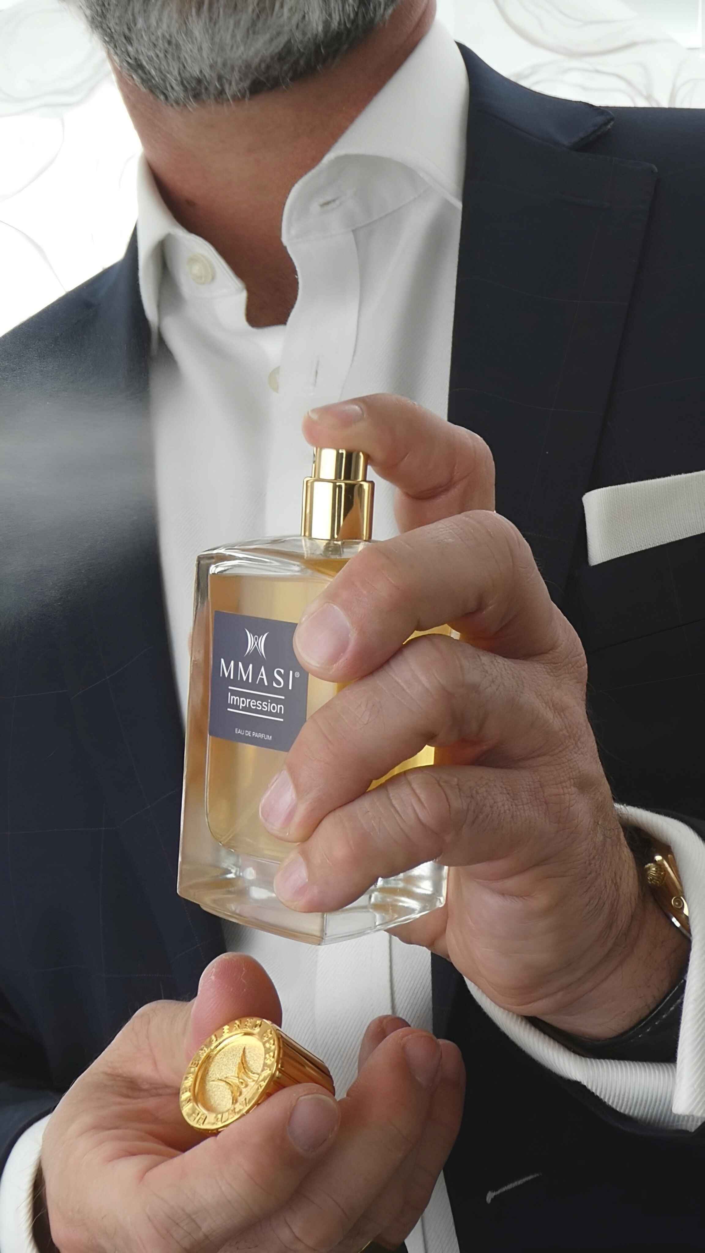 A man in a suit and white shirt holding a bottle of Mmasi Impression Eau de Parfum with his fingers on the spray nozzle, and holding the bottles gold cap on his right hand.